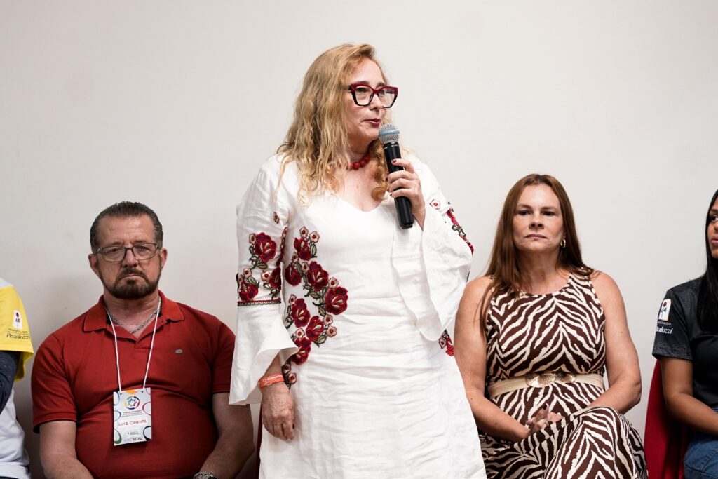 Mesa de abertura com participantes sentados diante de um palco de cor clara e paredes brancas. Fala ao microfone a presidente da Fenapestalozzi, Ester Pacheco, uma mulher loira de vestido branco.