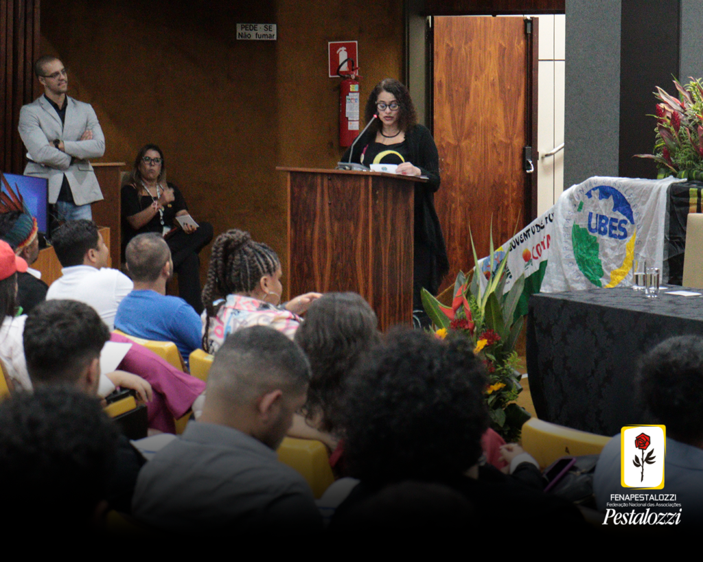 A ministra de Estado da Ciência, Tecnologia e Inovação, Luciana Santos, fala ao público diante de um púlpito de cor marrom, Ela veste um vestido preto, e usa óculos de armação escura. Seus cabelos são cacheados e estão na altura dos ombros, de cor castanha. 