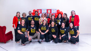 Equipe organizadora e presidentes da Fenapestalozzi. Há uma fila de pesoas em pé, e outra fila agachada, à frente. As pessoas vestem uma camiseta preta com os dizeres "#Rumo aos 100 anos Pestalozzi no Brasil". Atrás do grupo há uma parede de cor bege e um banner com a marca da organização nas cores vermelha e branca. 