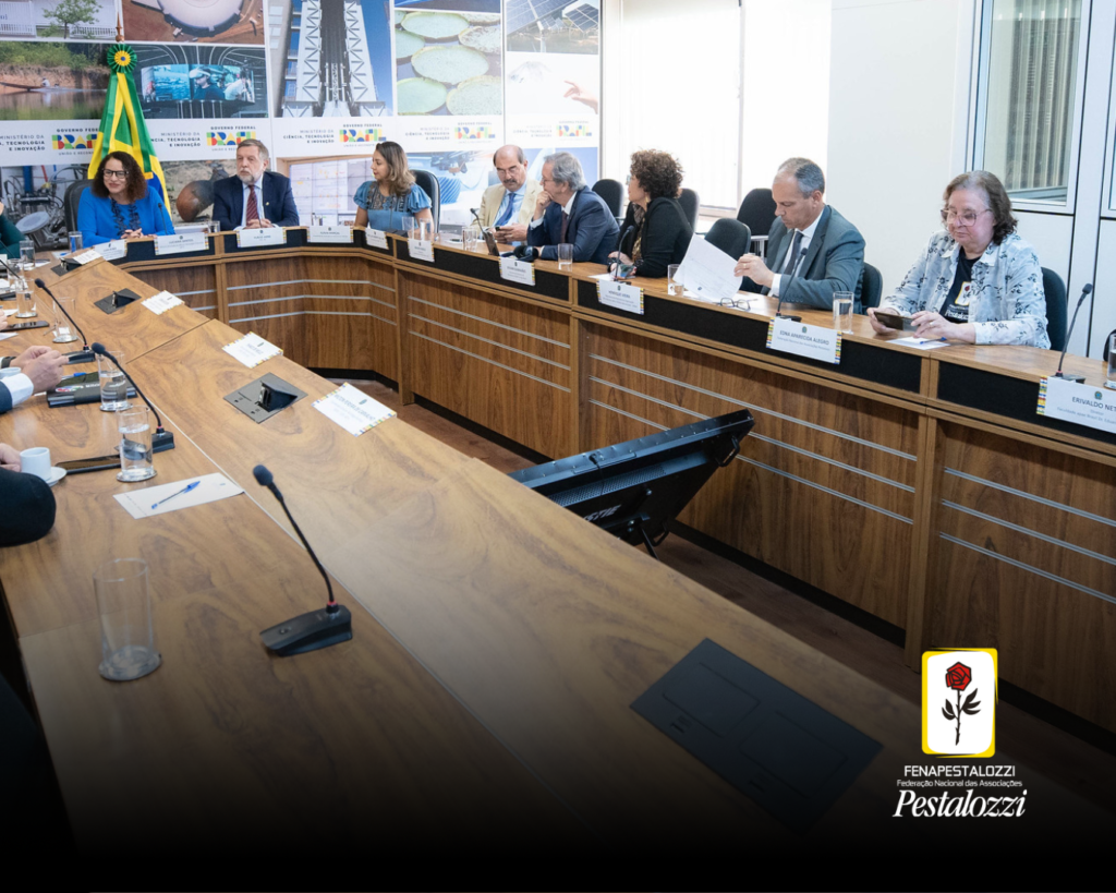 Mesa em formato de U, cor caramelo. As pessoas representantes da sociedade civil e governo estão ao derredor da mesa. Ao fundo da sala há fotografias e quadros pendurados em uma parede branca.