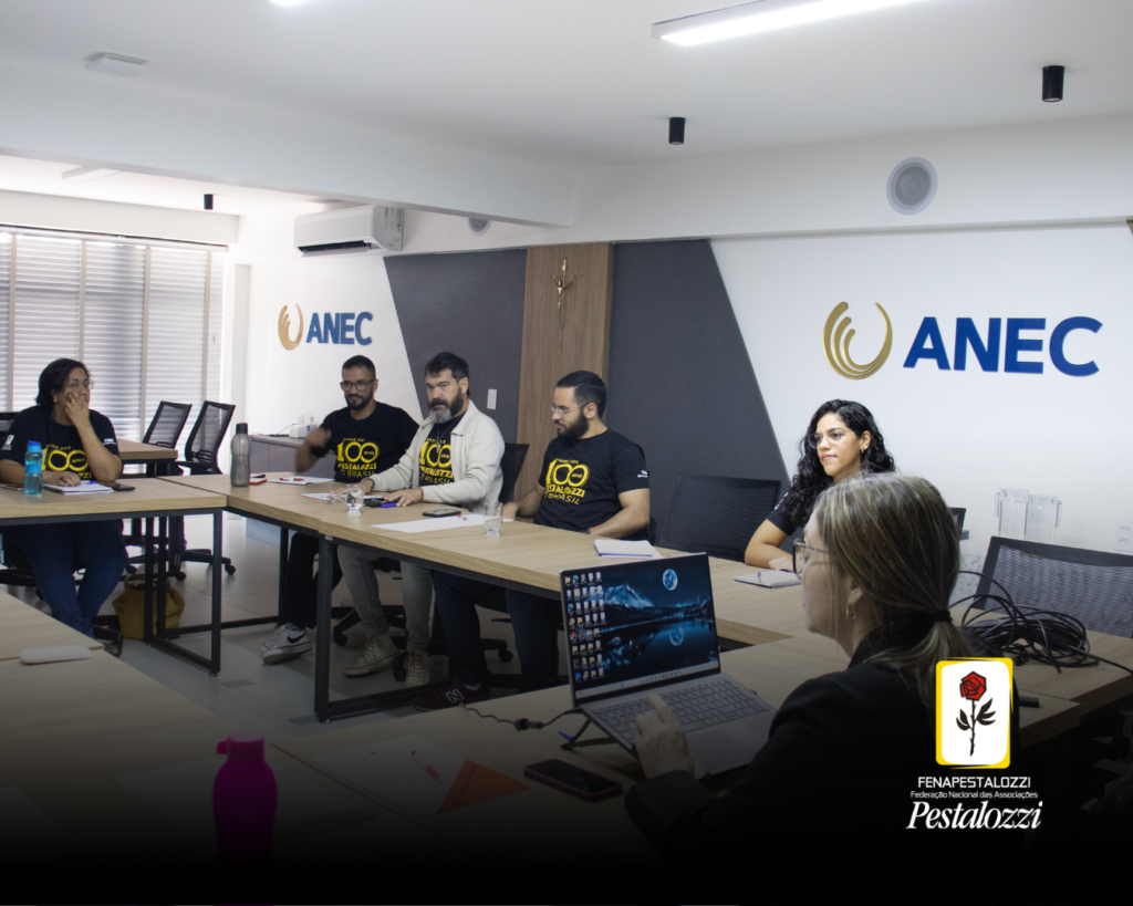 Profissionais da Fenapestalozzi estão sentados em formato de U, diante de uma mesa de cor amarela. Eles estão sentados e olhando para a frente. A ministrante da capacitação, Rozângela, uma mulher branca de cabelos claros, está de costas para a câmera e de frente para os participantes. Atrás dos profissionais, h[á uma parede de cor clara co a sigla ANEC na cor azul.