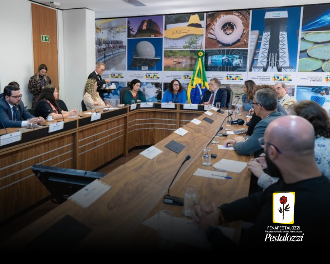 Mesa em formato de U, cor caramelo. As pessoas representantes da sociedade civil e governo estão ao derredor da mesa. Ao fundo da sala há fotografias e quadros pendurados em uma parede branca.