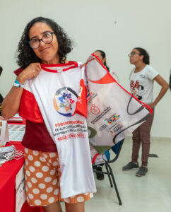 Autodefensora Nacional da região nordeste, Maria da Conceição. Maria é uma mulher parda, de cabelos castanhos na altura dos ombros. Ela segura uma camiseta branca com mangas vermelhas. Há pessoas não identificadas ao fundo. 