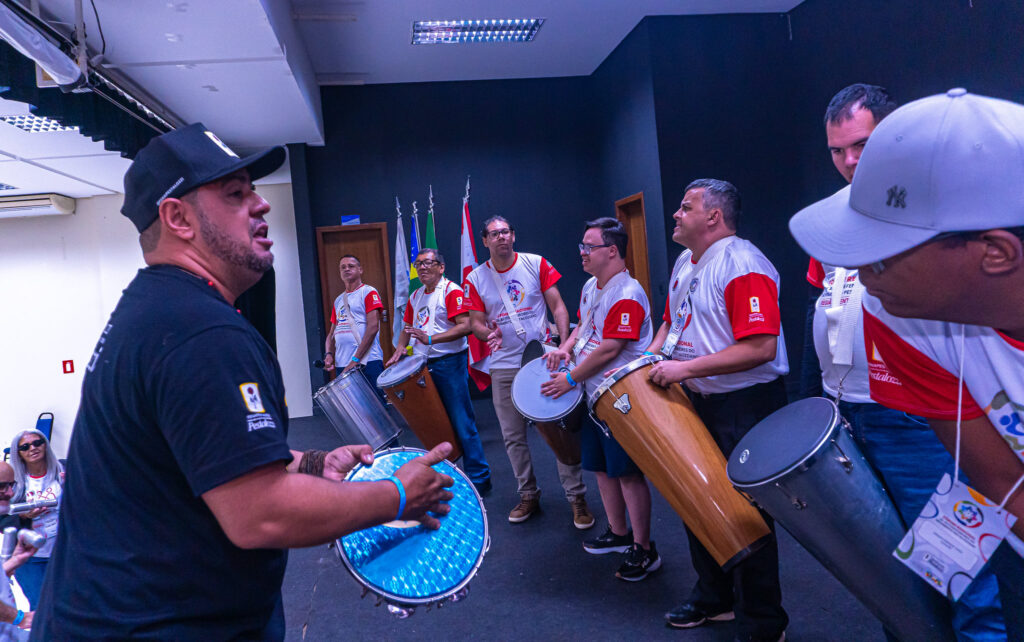 Grupo de autodefensores vestem uma camiseta branca com mangas vermelhas. Eles estão em fila no palco preto, e estão de frente para o músico Kerby Torres. Ele veste uma camiseta preta e um vone preto. Todos estão portando instrumentos musicais de percussão. 