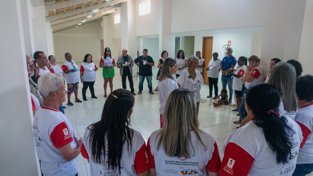 Grupo de pessoas se organizam em círculo. Elas vestem uma camiseta branca com mangas vermellhas. Ao centro, as duas coordenadoras de Família, Cícera Lopes e Antonieta Bonifácio. 