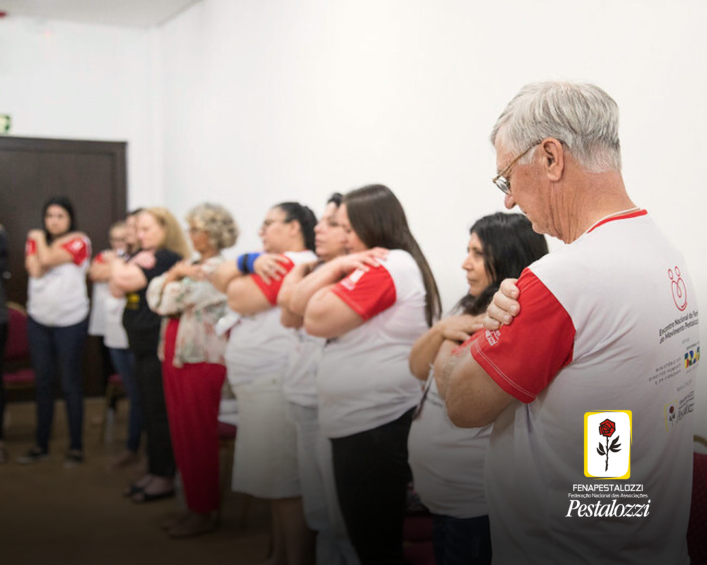 Foto de um grupo de pessoas em perspectivam que simulam um autoabraço. Elas estão em uma sala de cor branca e usam uma camiseta branca com mangas vermelhas. 