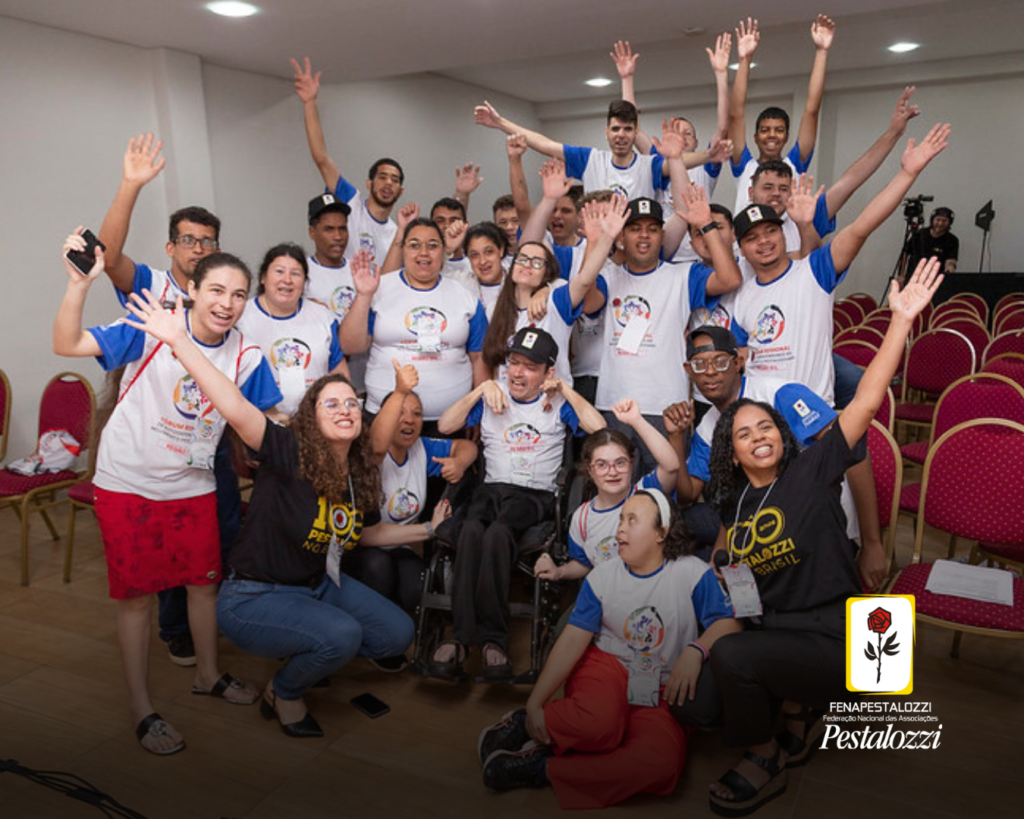 Grupo de autodefensores jovens que sorriem e acenam para a câmera; eles vestem uma camiseta branca com mangas azuis. Estão acompanhados das coordenadoras Nacionais do Monpad, que usam uma camiseta preta com os dizeres: #Rumo aos 100 anos de Pestalozzi no Brasil. Eles estão em uma sala de paredes brancas e cadeiras vermelhas. 