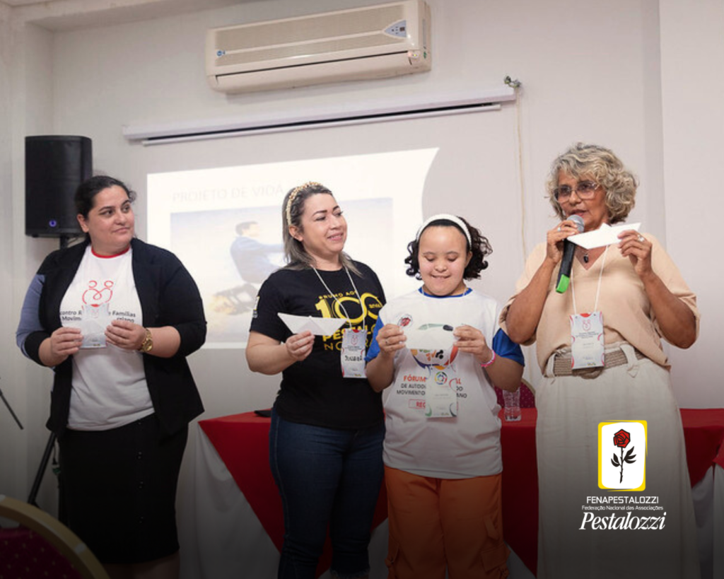 Quatro mulheres estão em pé e seguram um barco de papel, com escritos sobre seus objetivos de vida. A primeira da esquerda veste um casado preto e uma camiseta branca; a segunda mulher veste uma camiseta preta dos 100 anos do Movimento Pestalozziano; a terceira mulher usa uma camiseta branca com mangas azuis e está usando uma faixa branca na cabeça; e por último, a coordenadora Nacional de Famílias, que veste uma saia branca e uma blusa bege. 