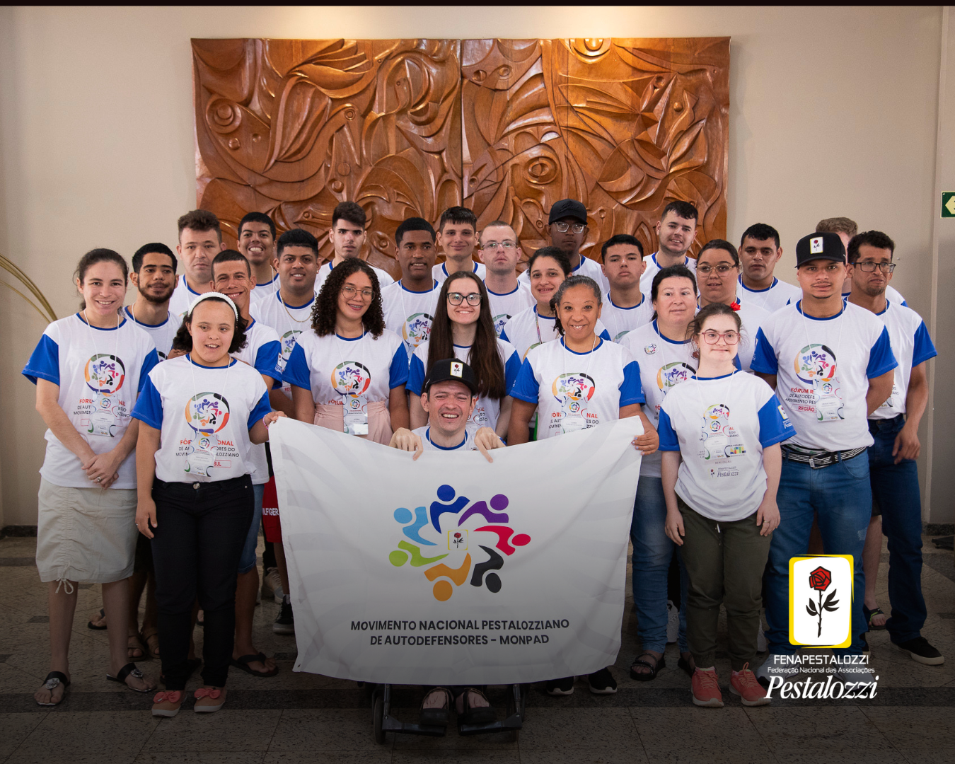 Grupo de autodefensores está de pé um ambiente de cor clara. Atrás deles uma parede de madeira. Trata-se de um grupo de jovens que vestem uma camiseta branca com mangas azuis. Mais adiante do grupo, um rapaz cadeirante porta uma bandeira do Monpad, de cor clara e símbolo colorido.