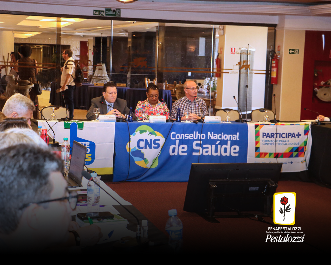 Foto em plano geral da mesa de discussão do CNS. Com três pessoas compondo a mesa diretora, à esquerda da imagem, um homem branco, de cabelos pretos e vestindo um terno cinza e camisa azul claro. Ao centro da mesa, uma mulher preta com cabelos pretos e trançados, utilizando um óculos de grau, vestindo uma blusa colorida estampada com flores. À direita da imagem, um homem branco, sem cabelos, utilizando óculos de grau e vestindo uma camisa quadriculada nas cores branco, marrom e preto. Estes estão diante de uma mesa coberta por bandeiras representando o Conselho Nacional de Saúde e Programas de Governo.