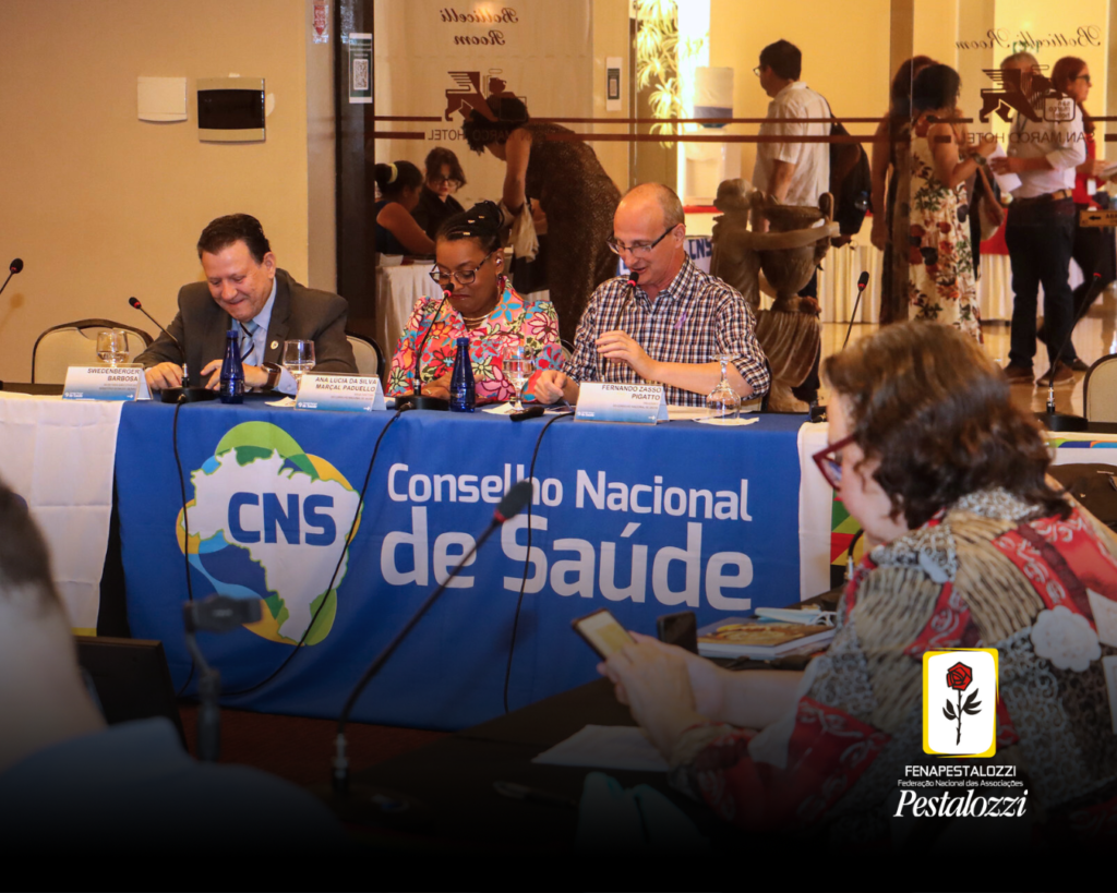 Foto em plano geral da mesa de discussão do CNS. Com três pessoas compondo a mesa diretora, à esquerda da imagem, um homem branco, de cabelos pretos e vestindo um terno cinza e camisa azul claro. Ao centro da mesa, uma mulher preta com cabelos pretos e trançados, utilizando um óculos de grau, vestindo uma blusa colorida estampada com flores. À direita da imagem, um homem branco, sem cabelos, utilizando óculos de grau e vestindo uma camisa quadriculada nas cores branco, marrom e preto. Ao canto direito da imagem, sentada à mesa, Edna Alegro, 2ª vice-presidente da Fenapestalozzi.Estes estão diante de uma mesa coberta por bandeiras representando o Conselho Nacional de Saúde e Programas de Governo.