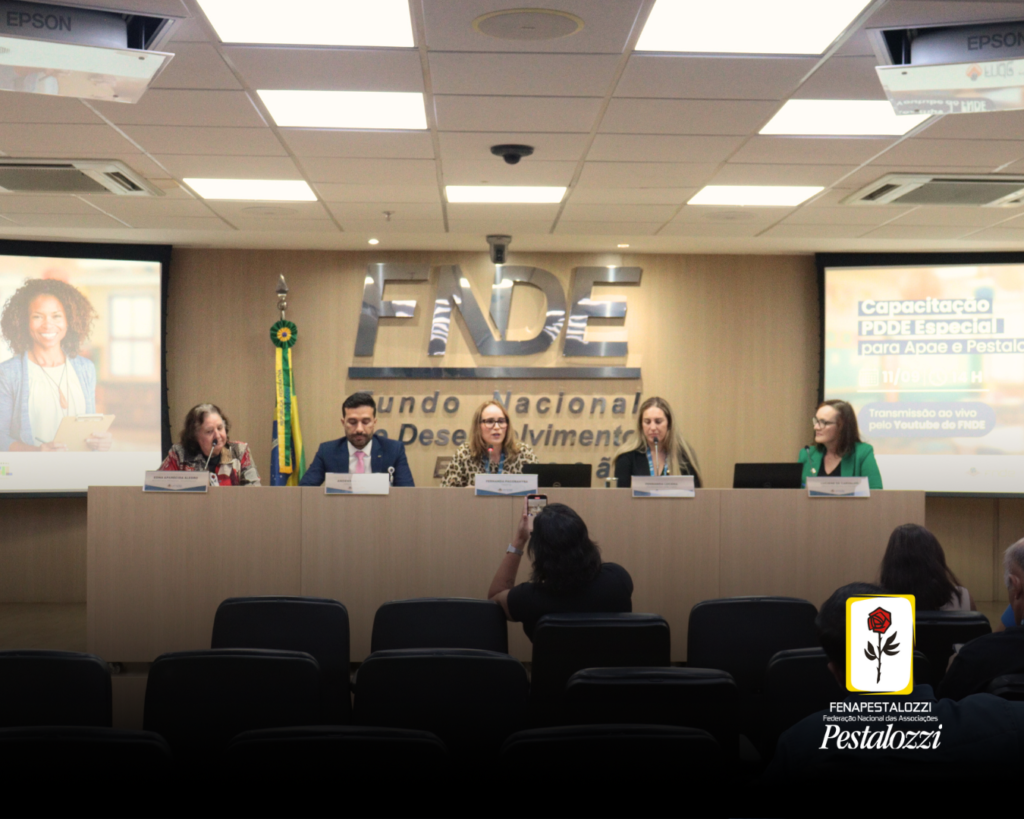 Foto em grupo dos participantes da mesa de abertura do webinário. Eles estão sentados em um púlpito de cor clara, e olham para o público que está na direção da foto. Ao fundo, há as letras prateadas "FNDE - fundo Nacional de Desenvolvimento da Educação", em um painel de cor clara também. A primeira pessoa à esquerda, trata-se da segunda vice-diretora da Fenapestalozzi, Edna Alegro, que é uma mulher branca, idosa, de cabelos castanhos na altura do ombro, e que usa óculos de armação escura. A segunda pessoa é o o diretor de Ações Educacionais, Anderson Santos, que veste um terno azul, de pele morena e cabelos curtos e castanhos; a terceira pessoa é a presidente do FNDE, Fernanda Pacobahyba, que é uma mulher branca, de cabelos loiros e que veste uma blusa com estampas de onça; a quarta pessoa é a coordenadora-geral do Desenvolvimento e Melhoria da Escola, Fernanda Lucena, que se trata de uma mulher branca, que veste um casaco preto; por último, a a gerente Institucional da Apae Brasil, Luciene de Carvalho, uma mulher branca, de cabelos castanhos, e que veste um casaco de cor verde.