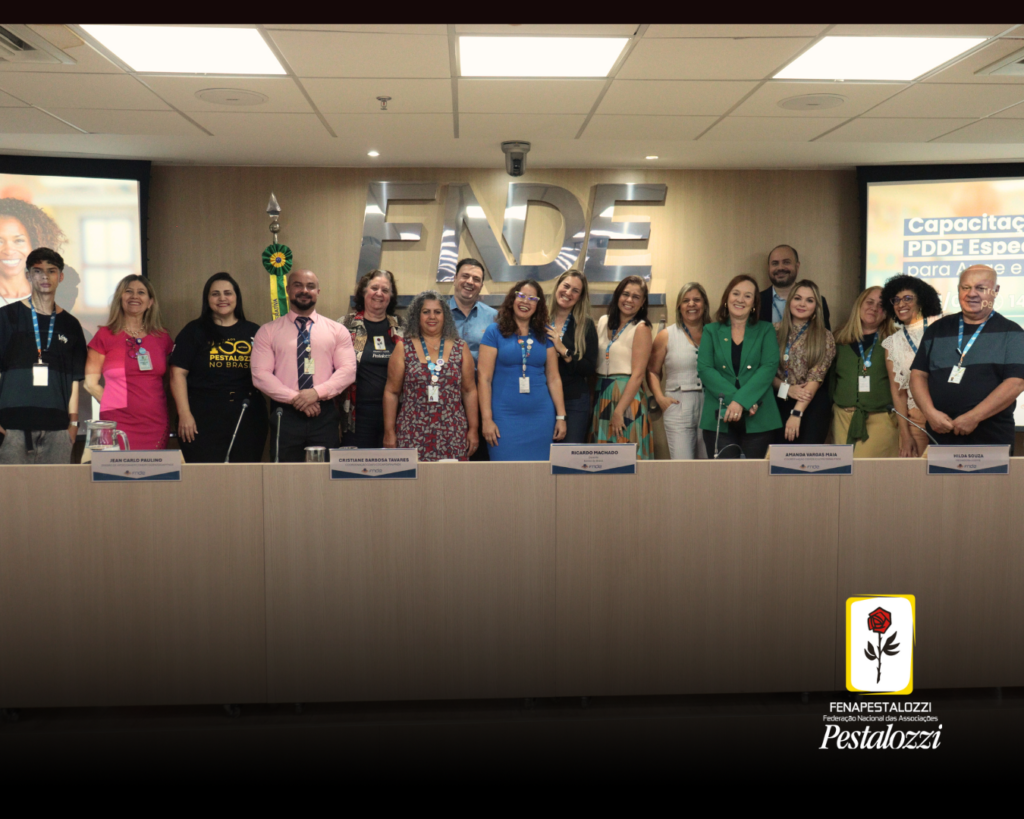 Foto em grupo dos participantes do webinários. A segunda vice-presidente da Fenapestalozzi, Edna Alegro, é a quinta pessoa da esquerda para a direita. O grupo de pessoas está diante do púlpito de cor clara, e, atrás deles, há o letreiro prateado com as sigla FNDE. 