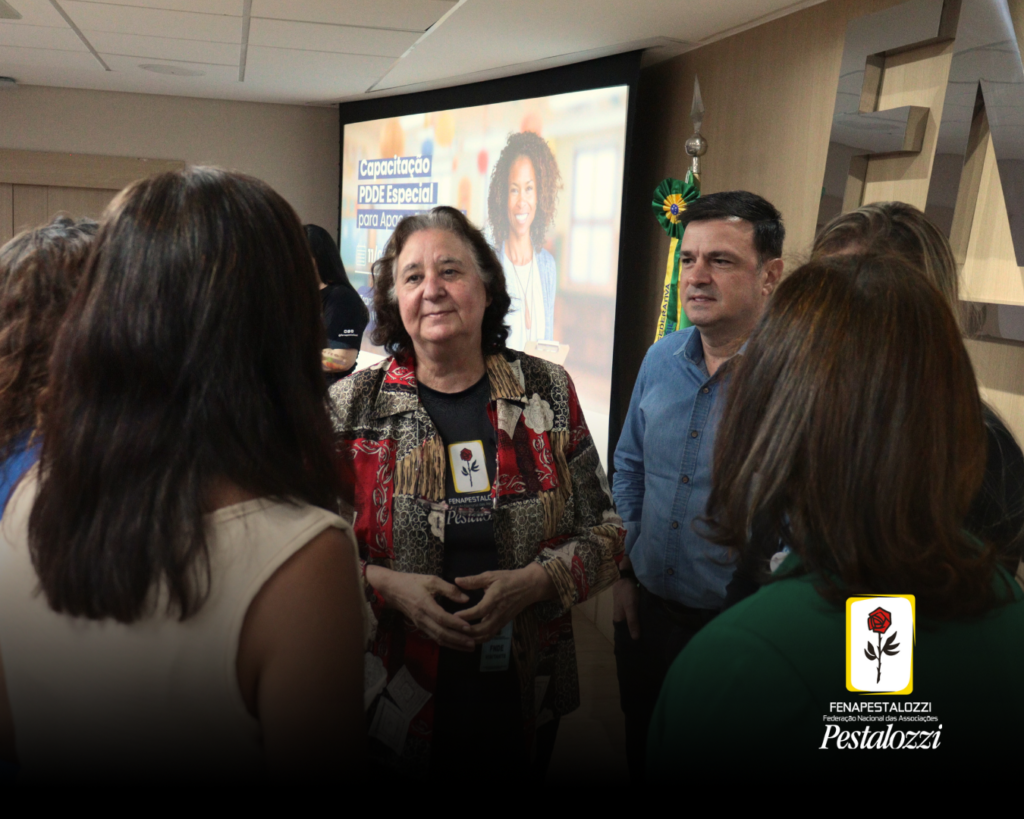 Grupo de pessoas conversa em semicírculo. Edna Alegro, segunda vice-presidente da Fenapesatalozzi está ao centrro. Ela é uma mulher idosa, branca, com cabelos curtos na altura do ombro. Veste uma camiseta preta com a marca da instituição ao centro e uma camisa estampada, ao seu lado esquerdo dela, está o Ricardo Américo de O. L. Machado, da diretoria de Governo, do Banco do Brasil. 