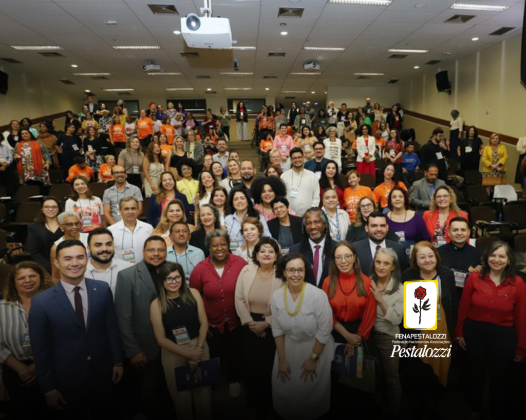 Foto em grupo em um auditório de paredes claras. As pessoas estão de pé e olham para a câmera. Na parte inferior da imagem, um efeito de leve escurecimento, e à direita, a assinatura completa da Federação Nacional das Associações Pestalozzi, Fenapeslozzi, com o ícone da rosa pestalozzina: botão de flor rosa na cor vermelha, caule, folhas e contornos na cor preta. O botão está envolvido em um moldura a amarela com fundo na cor branca.