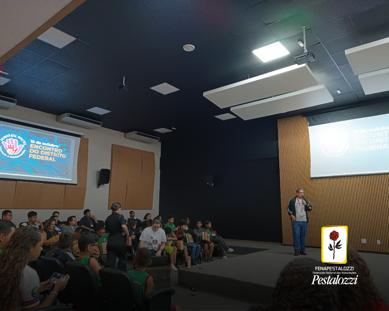 Fotomontagem. Há a foto de um auditório arena com algumas pessoas sentadas diante de um homem que está de pé, em um palco, e que fala ao microfone. As paredes têm cor clara e há painéis de madeira ripados de cor clara. Há a projeção da marca do evento ao fundo. Assina a foto a marca completa da Fenapestalozzi com a rosa pestalozziana.