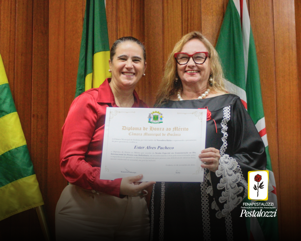 Fotomontagem. No canto inferior direito há a marca completa da Federação Nacional das Associações Pestalozzi com o ícone do Movimento Pestalozziano: um botão de roseira na cor vermelha, caule, folhas e contornos na cor preta. Este está envolvido em uma moldura amarela com fundo branco. Ao centro, a vereadora Kátia Maria e a presidente da Fenapestalozzi, Ester Pacheco. Mais à esquerda, Kátia, uma mulher branca de cabelos lisos e presos. Ela veste uma camisa vermelha e calça bege. Ao lado dela, mais à direita, Ester Pacheco: uma mulher branca de cabelos loiros e longos. Ela usa óculos de grau com armação grossa e vermelha e veste um vestido preto com detalhes em branco. Ambas seguram um certificado com texto não identificável e sorriem para o registro. Atrás delas, há um painel de madeira e as bandeiras do Brasil, de Goiás e de Goiânia.