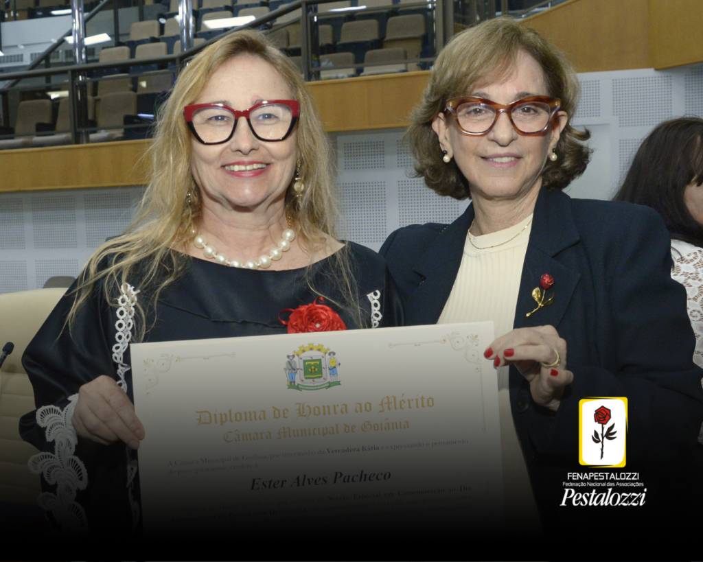 Fotomontagem. No canto inferior direito há a marca completa da Federação Nacional das Associações Pestalozzi com o ícone do Movimento Pestalozziano: um botão de roseira na cor vermelha, caule, folhas e contornos na cor preta. Este está envolvido em uma moldura amarela com fundo branco. Ao centro, foto em plano médio da Ester e da Carminha, presidente e vice-presidente da Fenapestalozzi. mais à esquerda está a Ester: ela é mulher branca de cabelos loiros e longos, usa óculos de grau com armação grossa e vermelha e veste um vestido preto com detalhes em branco. Ao lado dela está a Carminha: mulher branca de cabelos loiros escuros na altura dos ombros que usa óculos de grau com armação grossa e vermelha. Ela veste uma blusa bege e um blazer preto. Elas seguram o certificado da menção honrosa e sorriem para o registro. Ao fundo delas, há cadeiras de cor clara do Plenário da Casa.