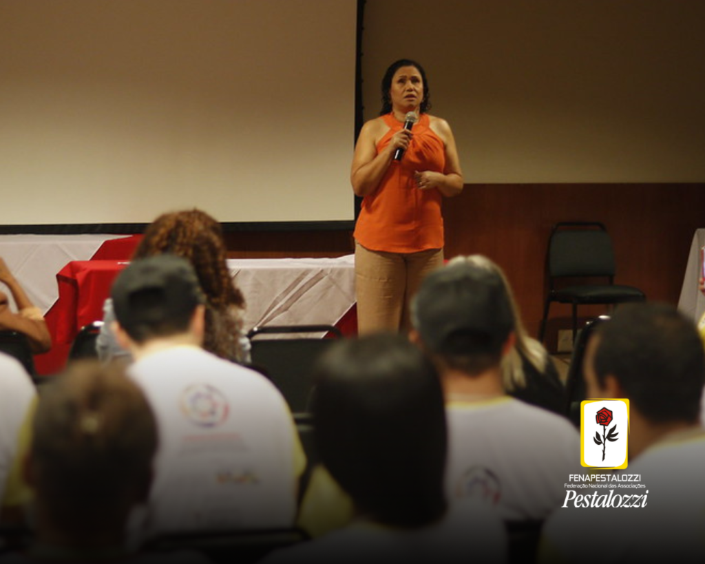 Fotomontagem. No canto inferior direito há a marca completa da Federação Nacional das Associações Pestalozzi com o ícone do Movimento Pestalozziano: um botão de roseira na cor vermelha, caule, folhas e contornos na cor preta. Este está envolvido em uma moldura amarela com fundo branco. Ao centro, foto de um auditório de paredes claras. Há pessoas que estão de costas para a fotografia e de frente para a palestrante. Esta é uma mulher parda de cabelos cacheados e curtos. Ela veste uma blusa alaranjada e uma calça de cor bege, e fala ao microfone. O auditório possui paredes claras.