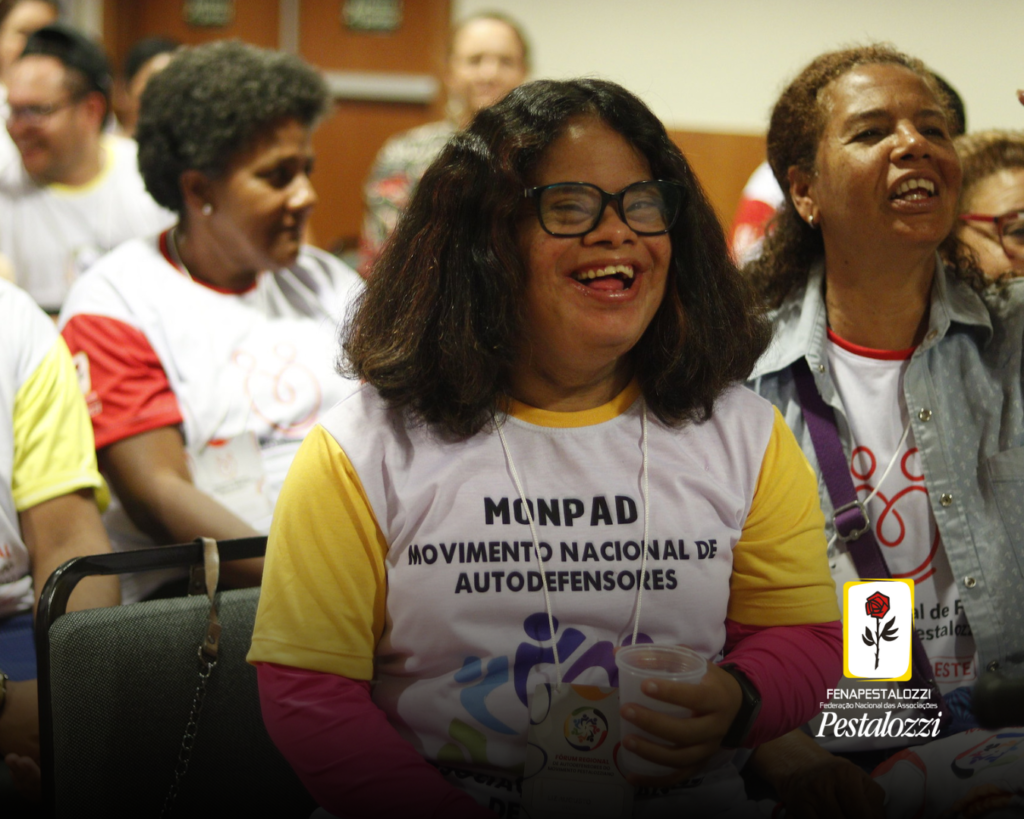 Fotomontagem. No canto inferior direito há a marca completa da Federação Nacional das Associações Pestalozzi com o ícone do Movimento Pestalozziano: um botão de roseira na cor vermelha, caule, folhas e contornos na cor preta. Este está envolvido em uma moldura amarela com fundo branco. Ao centro, foto em plano médio de uma menina com Síndrome de Down negra, de cabelos cacheados e óculos de grau com armação grossa e preta. Ela está sentada em um auditório de paredes claras e há pessoas não identificáveis atrás dela. 