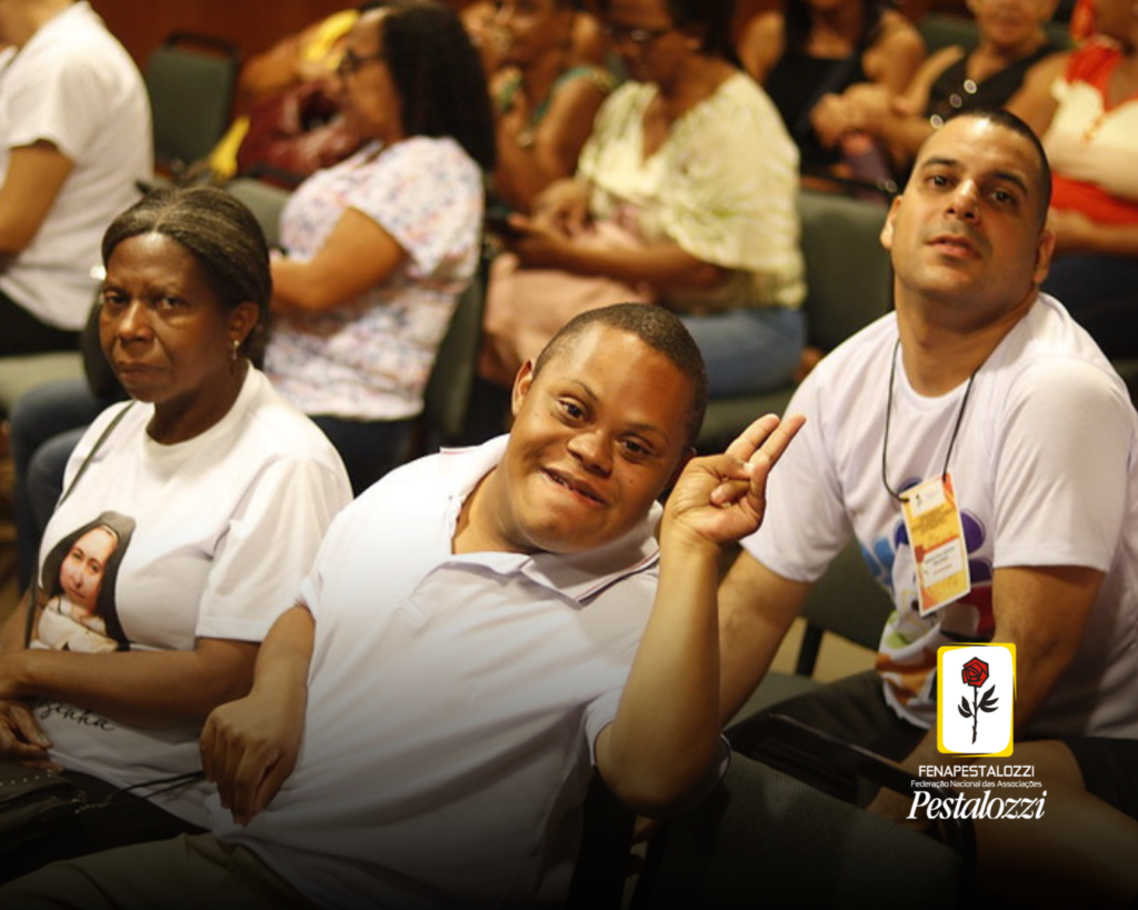 Fotomontagem. No canto inferior direito há a marca completa da Federação Nacional das Associações Pestalozzi com o ícone do Movimento Pestalozziano: um botão de roseira na cor vermelha, caule, folhas e contornos na cor preta. Este está envolvido em uma moldura amarela com fundo branco. ao centro, foto em plano médio de um rapaz cadeirante, negro, com Síndrome de Down que sorri e acena para a foto. Ao lado dele, à esquerda da foto, há uma mulher negra que olha serena para a câmera, e do outro lado, à direita, há um rapaz pardo de cabelos raspados que olha para a lente com sorriso discreto. Atrás deles há pessoas sentadas não identificáveis e uma sala de paredes claras. 