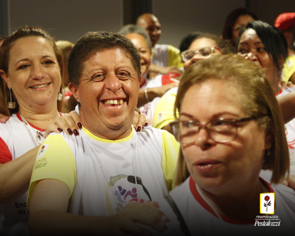 Fotomontagem. No canto inferior direito há a marca completa da Federação Nacional das Associações Pestalozzi com o ícone do Movimento Pestalozziano: um botão de roseira na cor vermelha, caule, folhas e contornos na cor preta. Este está envolvido em uma moldura amarela com fundo branco. Ao centro, participantes enfileirados com as mãos nos ombros uns dos outros sorriem e olham para a câmera. Eles estão em uma sala de paredes claras.