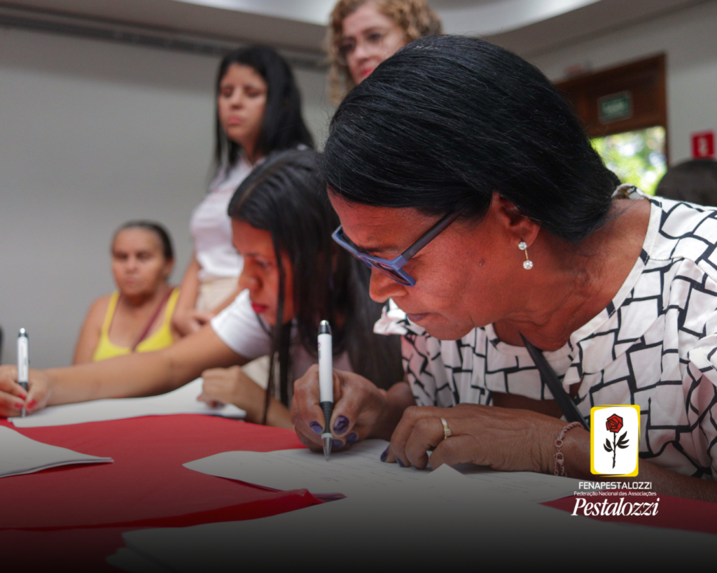 Fotomontagem. No canto inferior direito há a marca completa da Federação Nacional das Associações Pestalozzi com o ícone do Movimento Pestalozziano: um botão de roseira na cor vermelha, caule, folhas e contornos na cor preta. Este está envolvido em uma moldura amarela com fundo branco. Ao centro, foto em close de uma mulher negra de cabelos curtos e óculos de grau debruçada em uma mesa vermelha e branca, assinando papéis. Em perspectiva, há pessoas em pé, não identificáveis atrás dessa pessoa em primeiro plano.