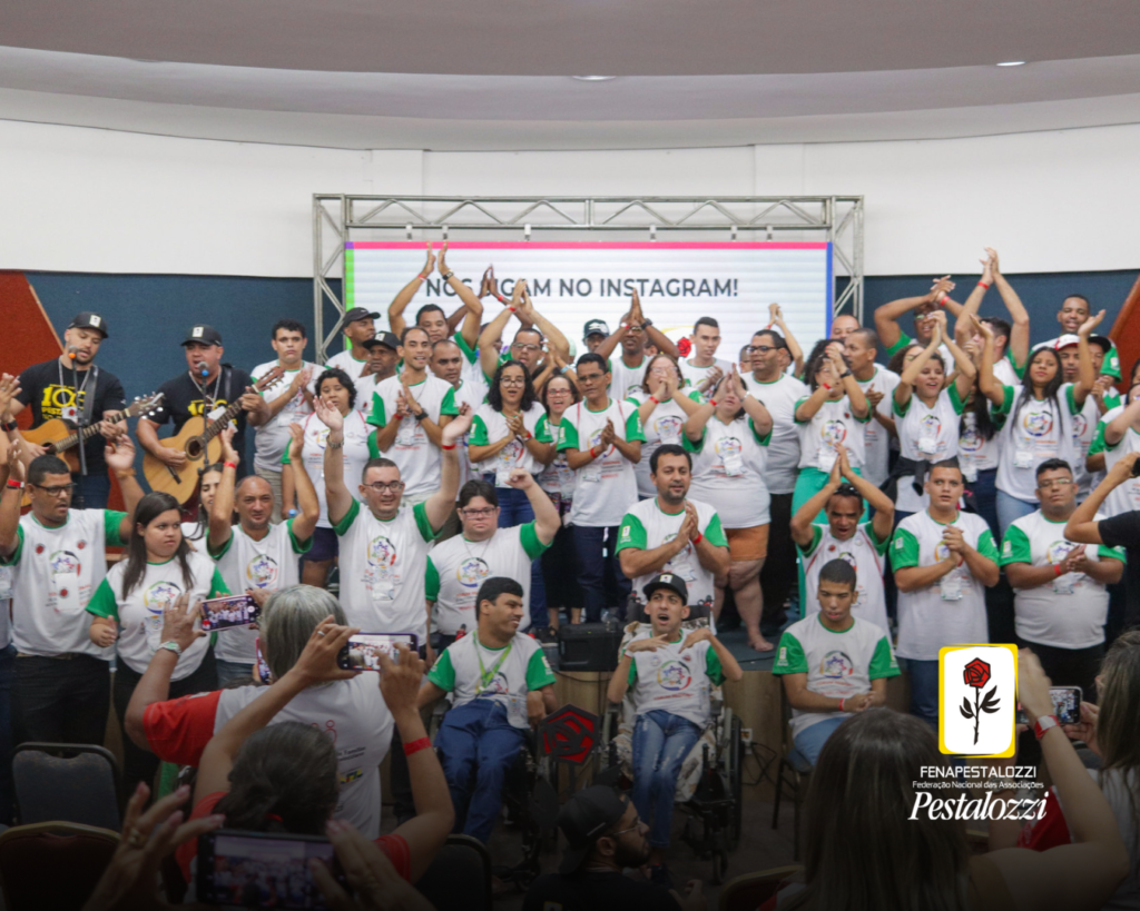 Fotomontagem. No canto inferior direito há a marca completa da Federação Nacional das Associações Pestalozzi com o ícone do Movimento Pestalozziano: um botão de roseira na cor vermelha, caule, folhas e contornos na cor preta. Este está envolvido em uma moldura amarela com fundo branco. Ao centro, foto em plano geral de autodefensores vestidos com a camiseta banca com mangas verdes. Eles estão diante de um painel de led em uma sala de paredes com metade azul e metade clara. Em contraplano há pessoas de costas, de frente para o grupo de autodefensores.