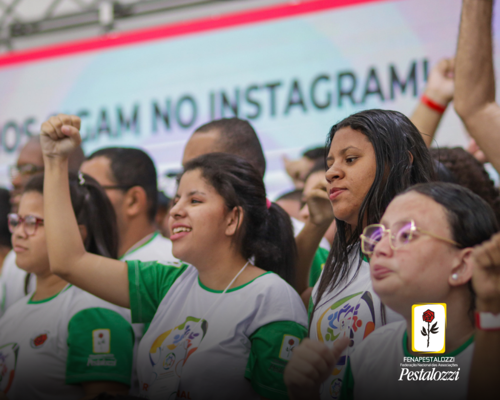 Fotomontagem. No canto inferior direito há a marca completa da Federação Nacional das Associações Pestalozzi com o ícone do Movimento Pestalozziano: um botão de roseira na cor vermelha, caule, folhas e contornos na cor preta. Este está envolvido em uma moldura amarela com fundo branco. Ao centro, foto em plano médio de um grupo de autodefensores que vestem uma camiseta branca e mangas verdes. Com destaque na foto, três mulheres que olham serenamente para frente, enquanto a câmera as registra lateralmente.