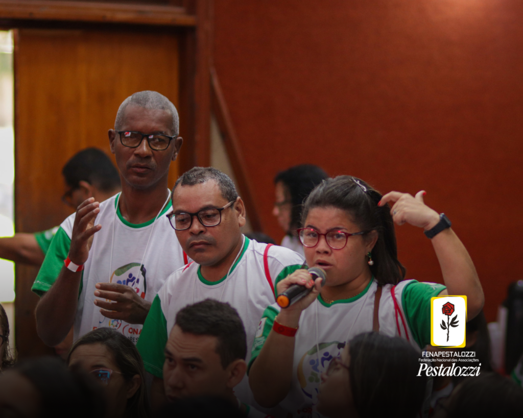 Fotomontagem. No canto inferior direito há a marca completa da Federação Nacional das Associações Pestalozzi com o ícone do Movimento Pestalozziano: um botão de roseira na cor vermelha, caule, folhas e contornos na cor preta. Este está envolvido em uma moldura amarela com fundo branco. Ao centro, foto em plano médio de três autodefensores que estão em pé diante de uma plateia. Eles vestem uma camiseta branca com mangas verdes. Da esquerda para direita, há um homem negro de cabelos grisalhos e curtos, óculos de grau de armação clara. Ao lado dele há um homem negro de cabelos curtos e óculos de grau de armação escura; por fim, à direita, há uma mulher negra de cabelos escuros na altura dos ombros e óculos de grau com armação vermelha que segura um microfone e gesticula.