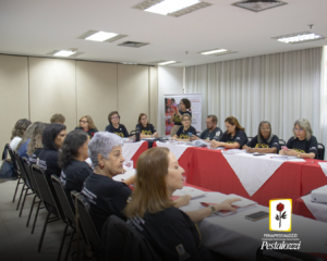 Fotomontagem com o ícone da Fenapestalozzi no canto inferior esquerdo: botão de roseira vermelha com caule, folhas e espinhos pretos. Este está envolvido em uma moldura amarela com fundo branco. A foto retrata uma mesa de reunião coberta por panos vermelho e branco. As pessoas estão dispostas em formato de "U", em uma sala de paredes e cortinas claras, vestindo uma camiseta preta com dizeres amarelos.
