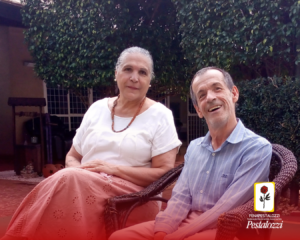 Fotomontagem em plano geral com a parte inferior da imagem com um elemento em degradê na cor vermelha e a assinatura da Fenapestalozzi com o ícone: botão de roseira vermelha com contornos, caule, espinhos e folhas na cor preta. Ele está envolto em uma moldura amarela com fundo branco. Ao centro está João e Lenilde, ambos sentados olhando para a câmera sorrindo. Lenilde é uma mulher branca, de cabelos grisalhos, que está vestindo uma blusa branca e uma saia rosa. João é um homem branco de cabelos raspados, que está de camisa de risca azul e calça branca. Completa a cena um jardim de folhes verde-escuras ao fundo.