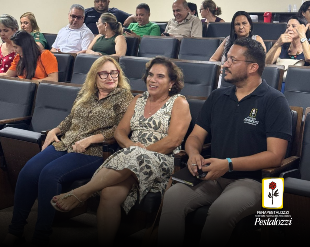Fotomontagem com a parte inferior ligeiramente escura e com a marca da Fenapestalozzi no canto inferior direito. Trata-se de um desenho de botão de roseira na cor vermelha com contornos, caule e espinhos na cor preta. Este está envolvido em uma moldura amarela com fundo branco. Ao centro, foto em primeiro plano de três pessoas que estão sentadas diante de um auditório de paredes claras e cadeiras escuras. As demais pessoas que compõe o auditório não são identificáveis. A primeira da esquerda trata-se de Ester, mulher branca, de cabelos loiros longos, óculos de grau com armação vermelha. Ela usa uma camisa de estampa de animal e calça jeans azul. Ao centro, Geli Sanches, mulher branca de cabelos na altura dos ombros que está vestindo um vestido branco com folhas verdes. À direita, Gustavo Almeida, homem pardo de cabelos castanhos curtos. Ele veste uma camiseta preta e calça cinza.