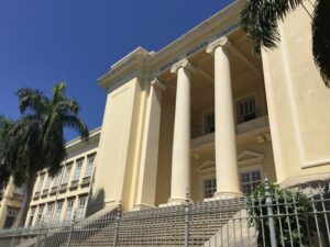 Foto em plano geral do Instituto Benjamin Constant. A imagem retrata um prédio de cor clara, janelas retangulares no estilo colonial e duas colunas redondas na parte da entrada do prédio, precedida por uma escadaria. O prédio parece cercado por grades e há palmeiras nas laterais. 