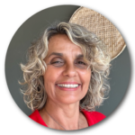 Retrato em close de Antonieta Bonifácio. Ela é uma mulher branca, de cabelos loiros e ondulados na altura dos ombros. Ela veste uma blusa vermelha e sorri para a foto. Atrás dela há uma peneira de palha na parede, que tem cor cinza. 