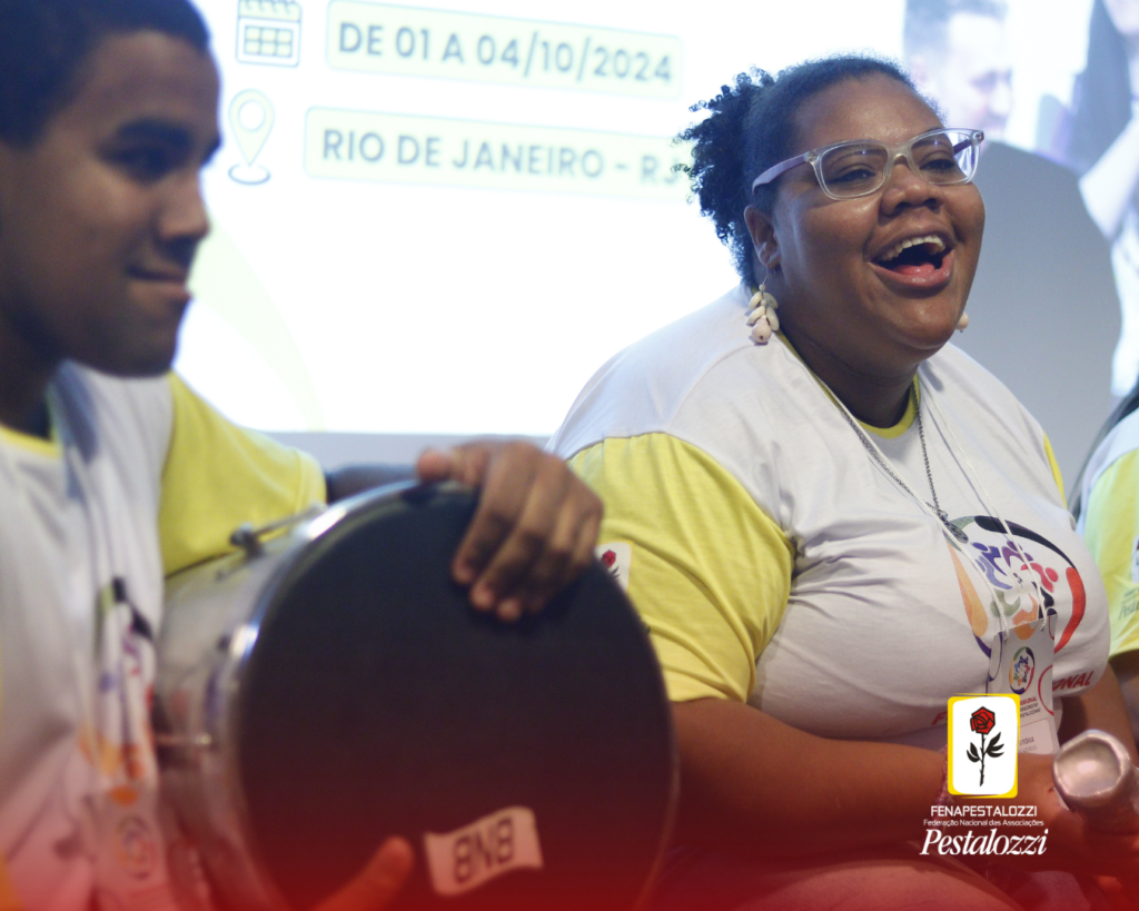 Fotomontagem: card com a parte inferior em degradê na cor vermelha, com a marca da Fenapestalozzi no canto inferior direito. Na parte central há a foto em plano médio de um rapaz e uma moça, ambos autodefensores, Ele, em primeiro plano, está com uma camiseta branca com mangas amarelas. É um rapaz negro de cabelos curtos que segura um instrumento de percussão. Ela, uma moça negra de cabeços crespos, presos. Está vestindo uma camiseta branca com mangas amarelas e óculos de grau com armação transparente. Ela segura um caxixi.