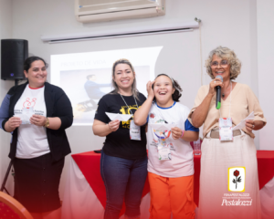 Fotomontagem: card com a parte inferior em degradê na cor vermelha, com a marca da Fenapestalozzi no canto inferior direito. Na parte central há a foto em plano médio de um rapaz e uma moça, ambos autodefensores, Ele, em primeiro plano, está com uma camiseta branca com mangas amarelas. É um rapaz negro de cabelos curtos que segura um instrumento de percussão. Ela, uma moça negra de cabeços crespos, presos. Está vestindo uma camiseta branca com mangas amarelas e óculos de grau com armação transparente. Ela segura um caxixi.