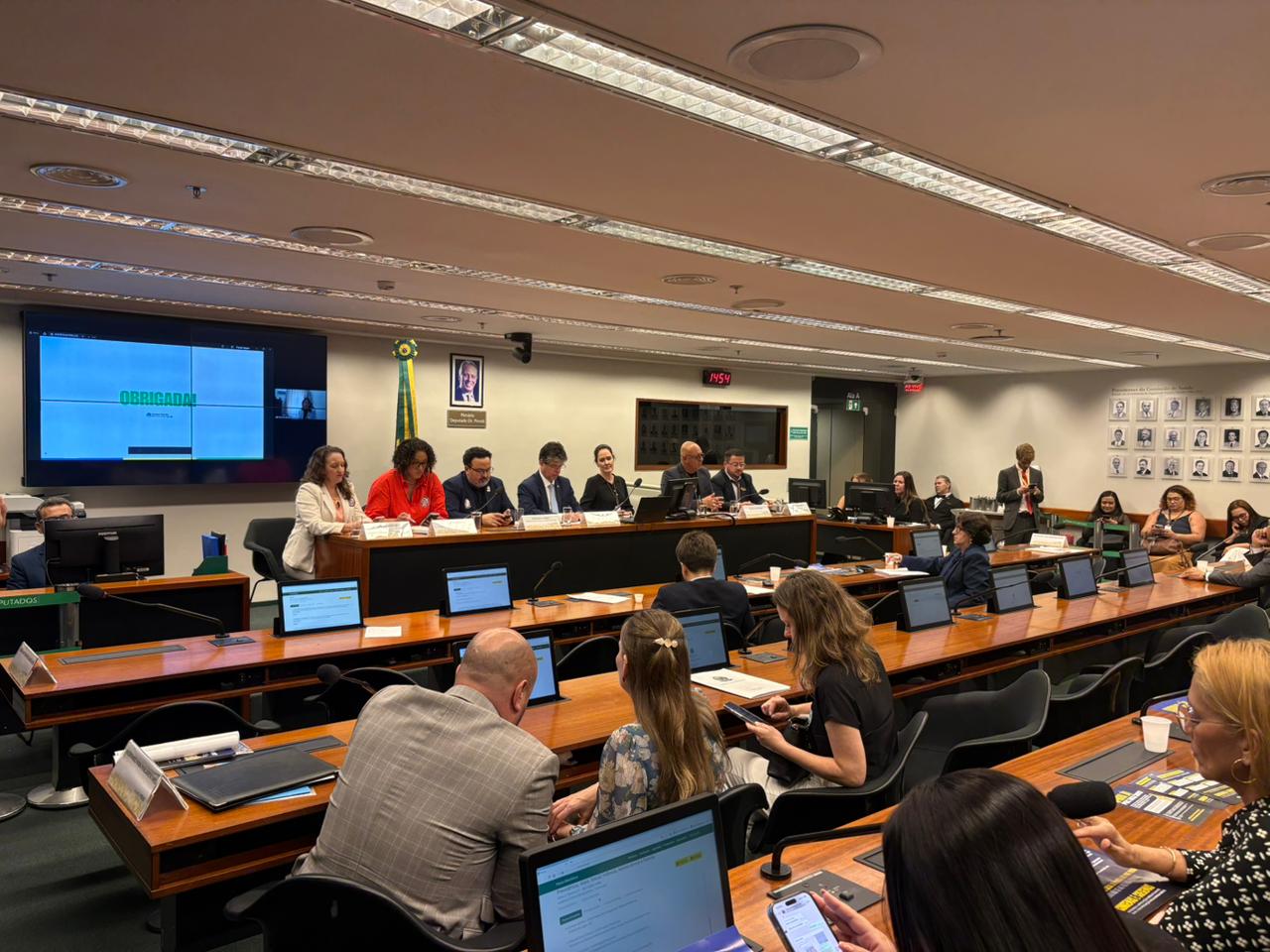 Uma foto de ângulo aberto de uma audiência em uma sala grande, no Congresso Nacional do Brasil, em Brasília, dada a decoração e o contexto.Primeiro Plano: Diversas pessoas sentadas em bancadas de madeira com computadores laptops à frente, voltadas para a frente da sala. Segundo Plano: Um grupo de palestrantes (aproximadamente 10 pessoas) está sentado em uma longa mesa elevada na frente da sala. Há um monitor grande à esquerda que exibe a palavra "OBRIGADA!" em letras verdes e brancas. Detalhes: A sala possui iluminação de teto, painéis de madeira e, no fundo, uma parede com retratos emoldurados. Uma pessoa está de pé à direita, falando com o grupo. A reunião parece ser formal, com a maioria dos participantes vestindo trajes sociais.