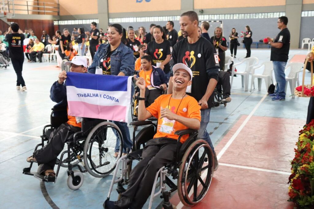Fotografia colorida em plano médio capturada em um ginásio poliesportivo. Em destaque, um jovem sorridente com deficiência física usa camiseta laranja e boné branco, sentado em uma cadeira de rodas que é conduzida por um homem negro com camiseta preta da Pestalozzi. Ao lado deles, outro jovem em cadeira de rodas segura com orgulho a bandeira do estado do Espírito Santo, que possui as cores azul, branco e rosa com a frase "Trabalha e Confia". Ao fundo, diversas pessoas em pé e em cadeiras de rodas participam do evento em um clima de celebração e alegria.