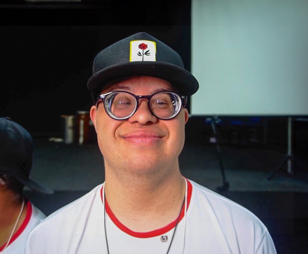 Fotografia em close-up de um homem jovem com síndrome de Down, sorrindo, usando óculos de lentes grossas, boné preto com o desenho de uma rosa e camiseta branca com gola vermelha.